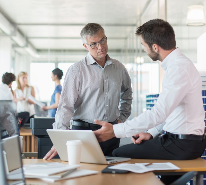 Two small business men standing in front of laptop while team meets in background